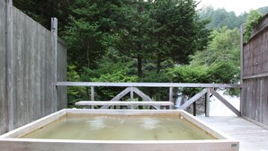 A wooden hot tub with a wooden deck and railing, surrounded by lush greenery.