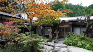 A traditional Japanese building with a wooden pathway leading to the entrance, surrounded by autumn foliage and greenery.