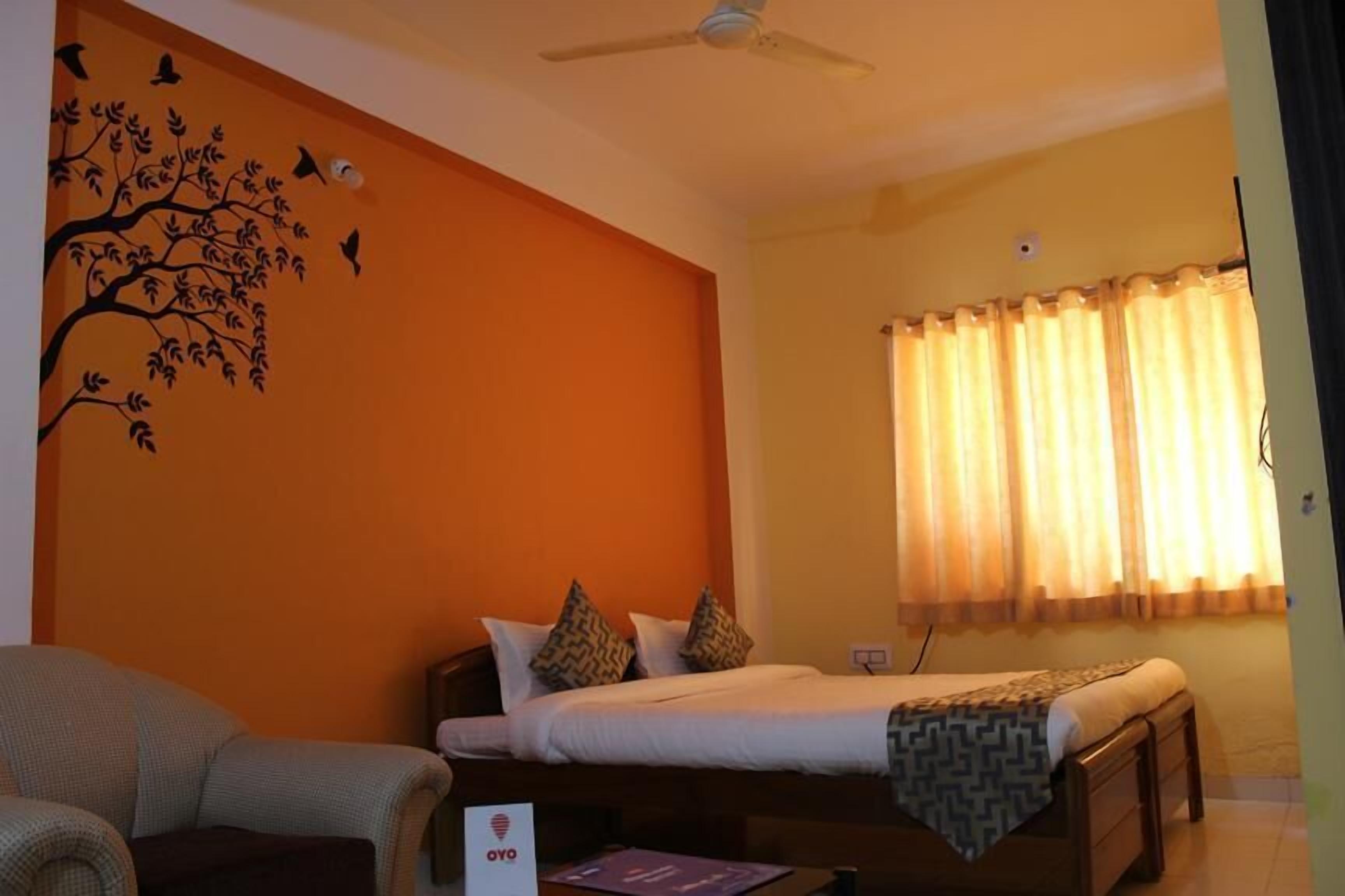 A hotel room with an orange accent wall, a bed with patterned pillows, a ceiling fan, and a window with curtains.