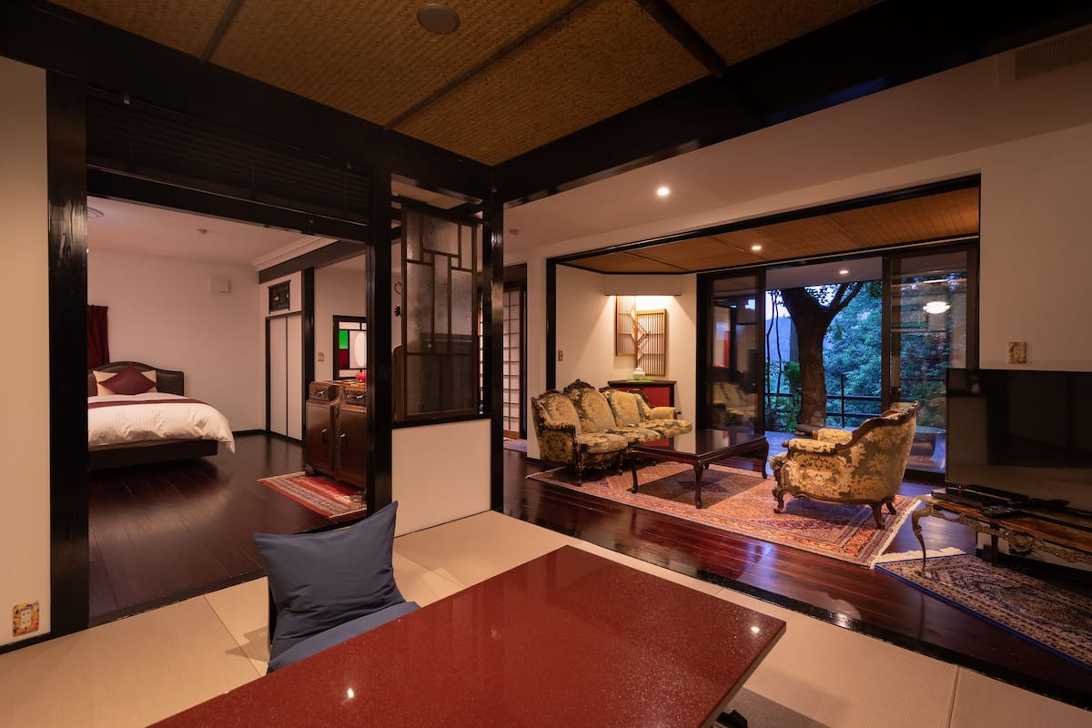 A modern hotel room with a large bed, a sitting area with a table and chairs, and a view of the outdoors through large windows.