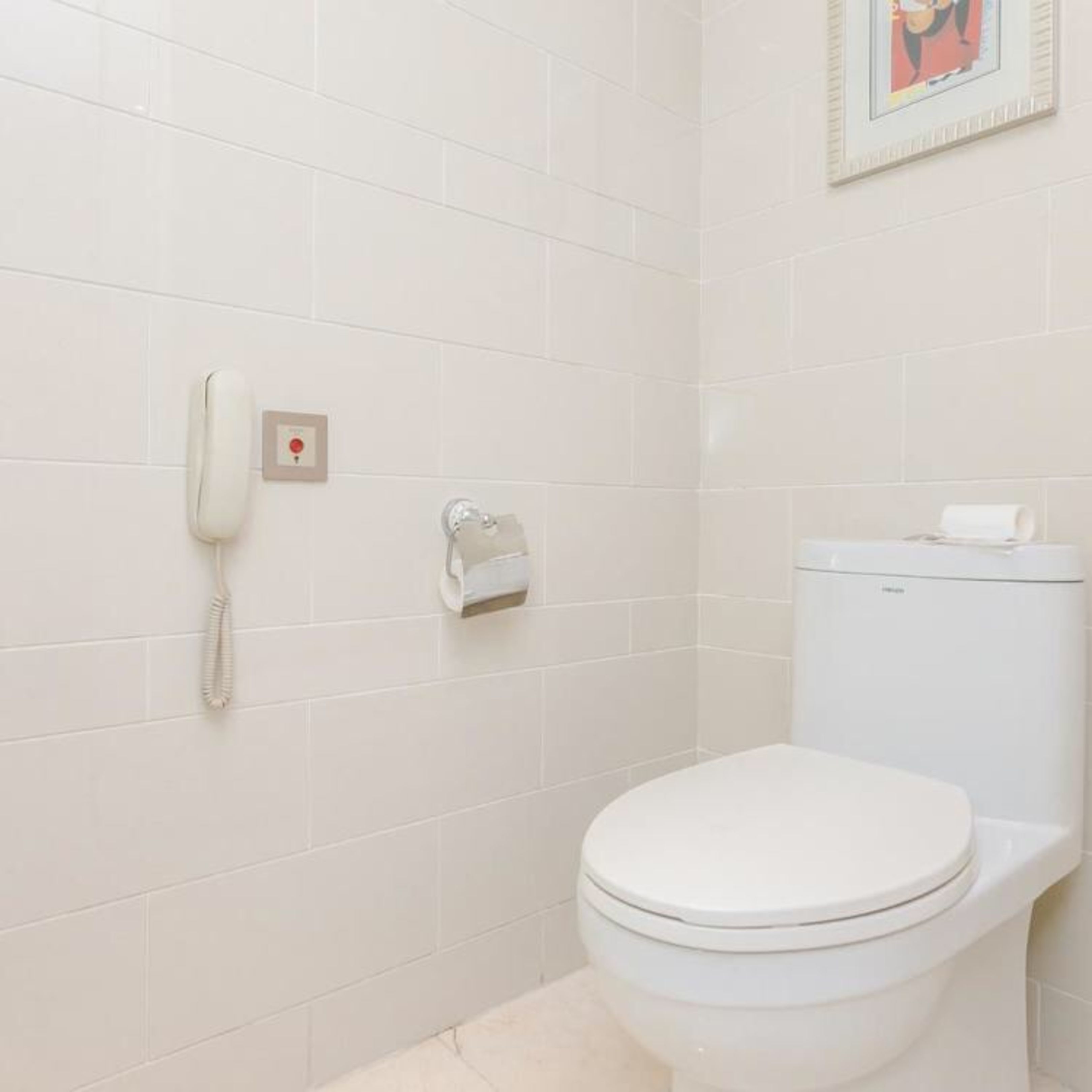 A bathroom with white tiles, a toilet, a toilet paper holder, and a wall-mounted telephone.