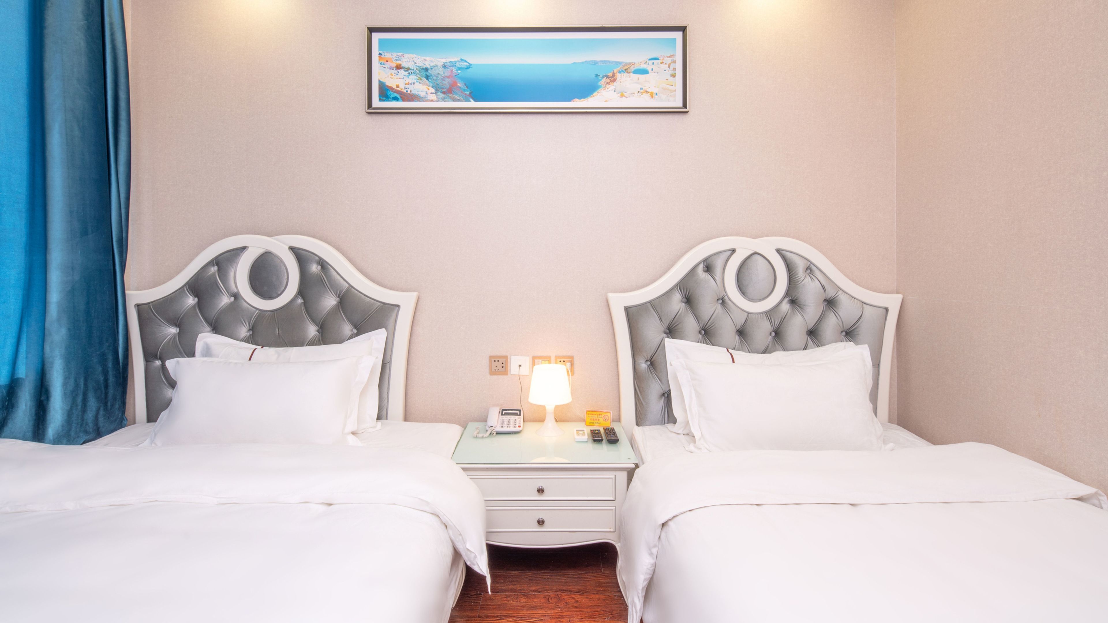 A hotel room with two beds, a nightstand with a lamp, a painting of a coastal landscape, and a blue curtain.