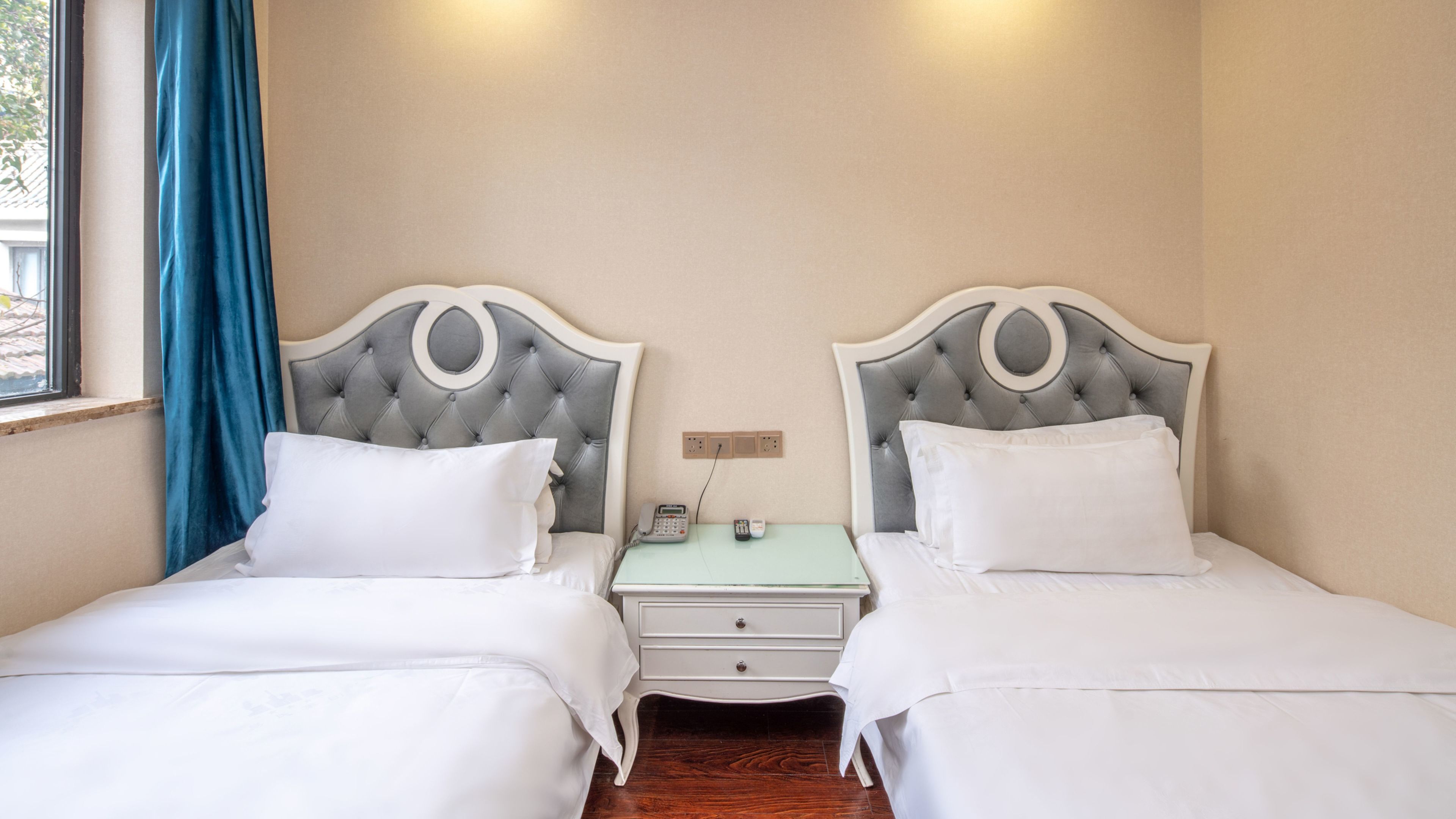 A double room with two beds, each with a tufted headboard, a bedside table with a telephone, and a wall socket.