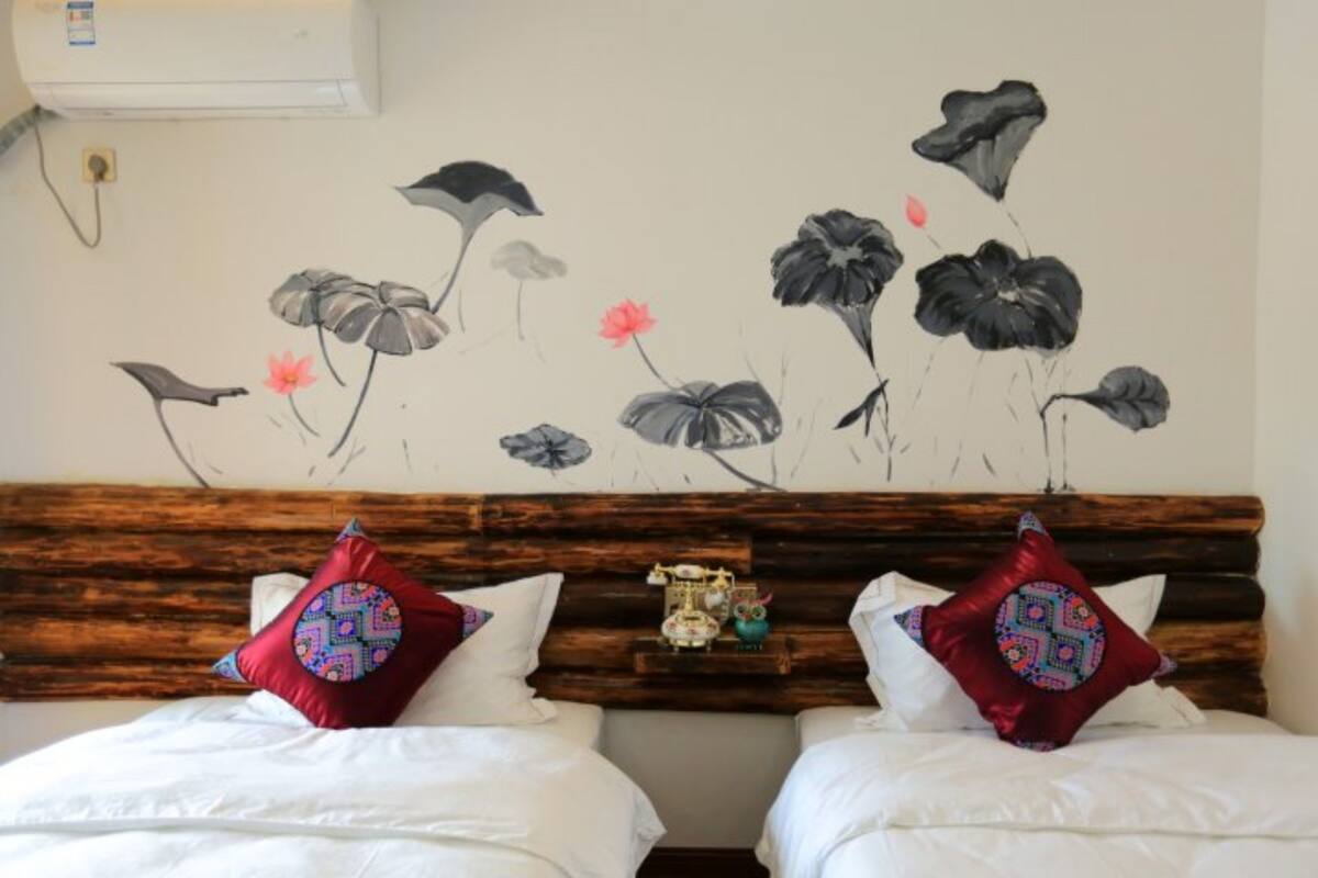 Quarto de hotel com duas camas, cada uma com roupa de cama branca e almofadas decorativas. Uma cabeceira de madeira separa as camas, e um mural na parede retrata flores e pássaros.