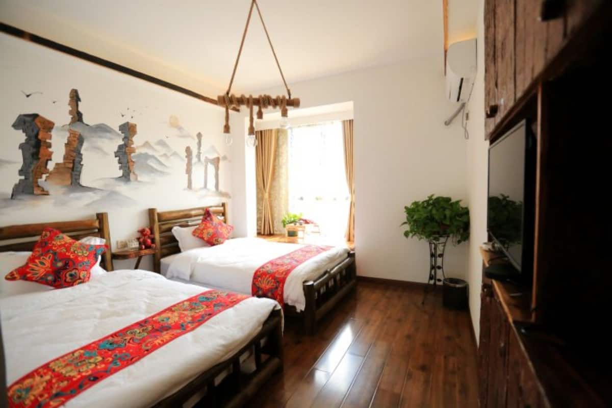 Quarto de hotel com duas camas, piso de madeira, uma janela grande com cortinas e um lustre.