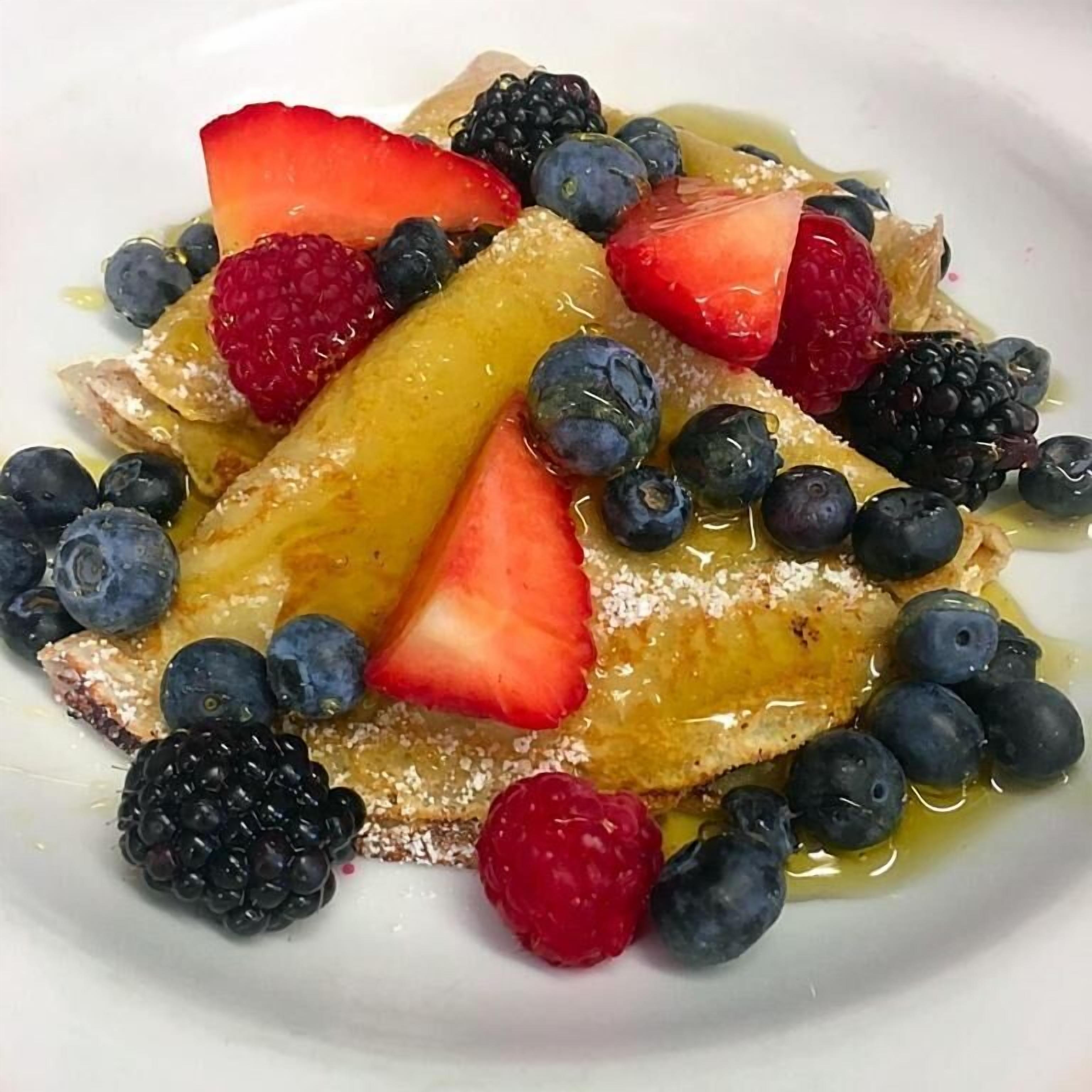 A plate of pancakes topped with strawberries, blueberries, raspberries, and blackberries, drizzled with syrup.