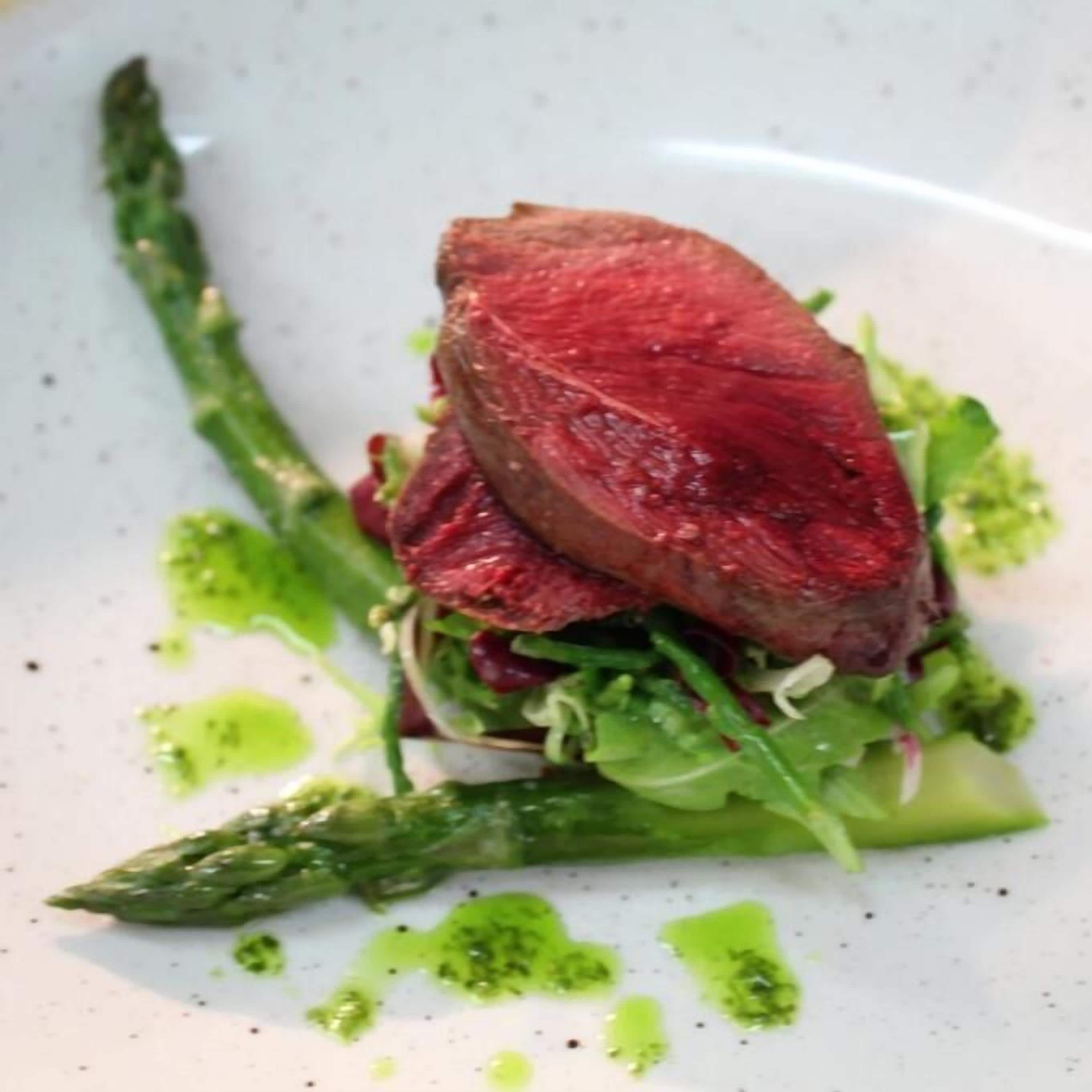 Een bord met een steak, asperges en groene bladgroenten.