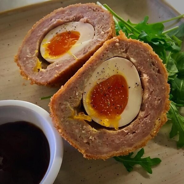 Dos mitades de huevo escocés con un huevo duro en el centro, servido en un plato con vegetales y un recipiente pequeño con salsa.