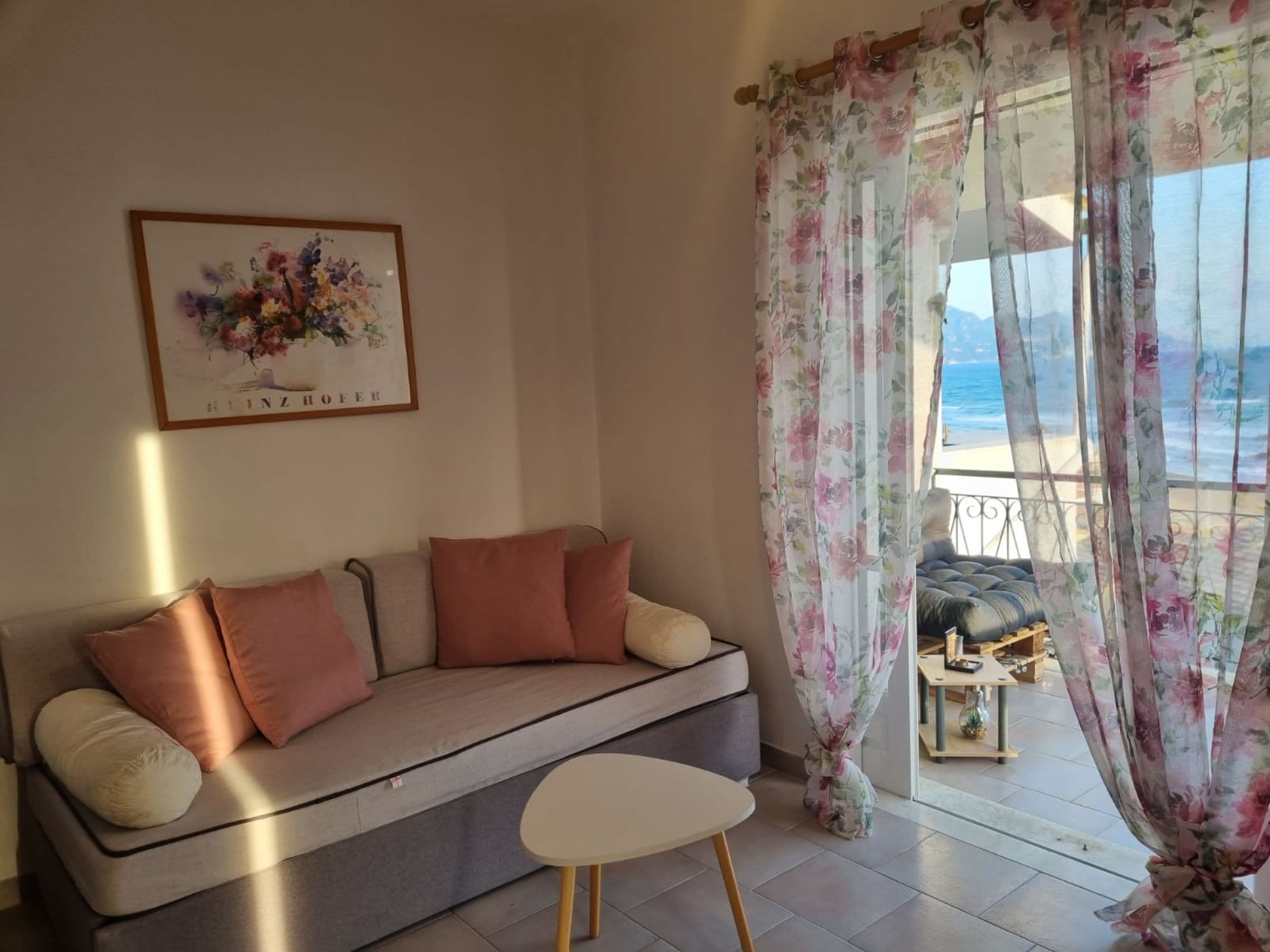 A living room with a sofa, a small table, and a view of the sea through a window with floral curtains.