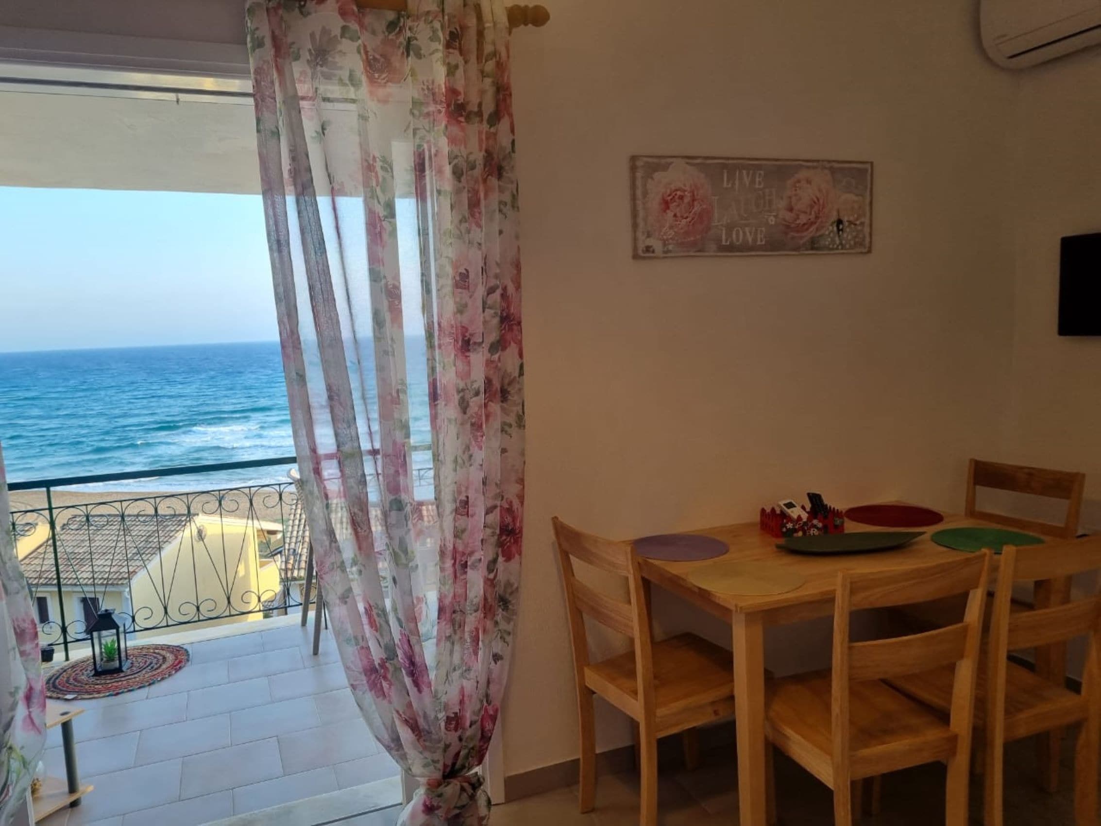 Un balcón con vista al mar, una mesa de madera con sillas y un cortinaje floral.