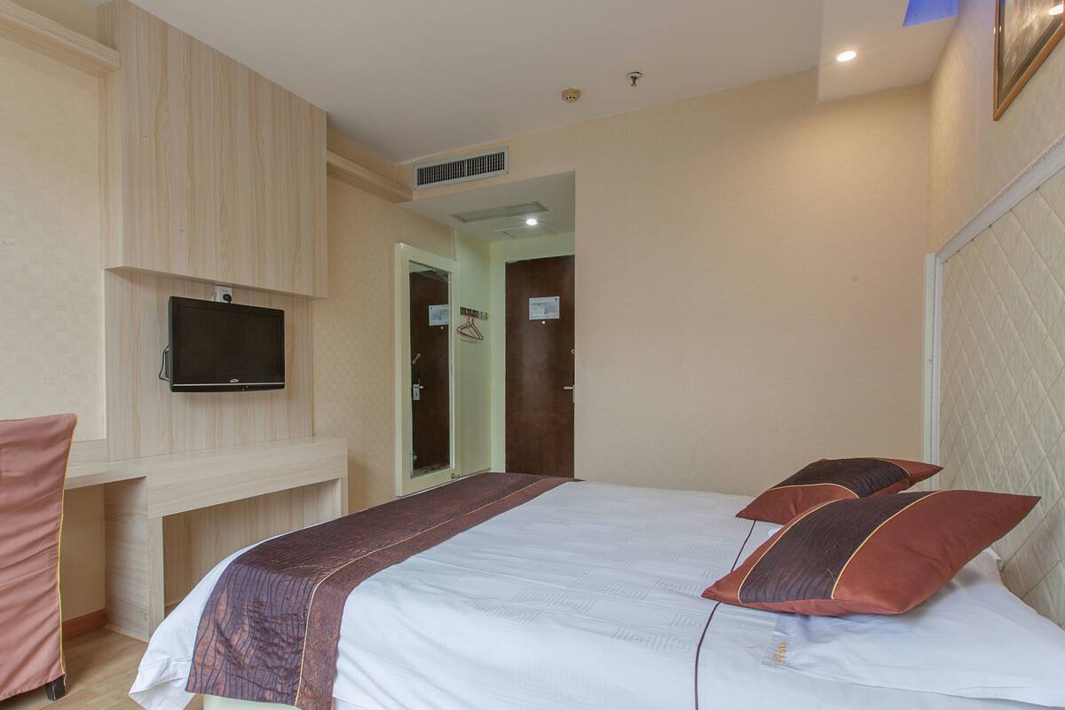 Ein Hotelzimmer mit einem Bett, einem an der Wand befestigten Fernseher, einem Spiegel und einer Tür zu einem anderen Raum.
