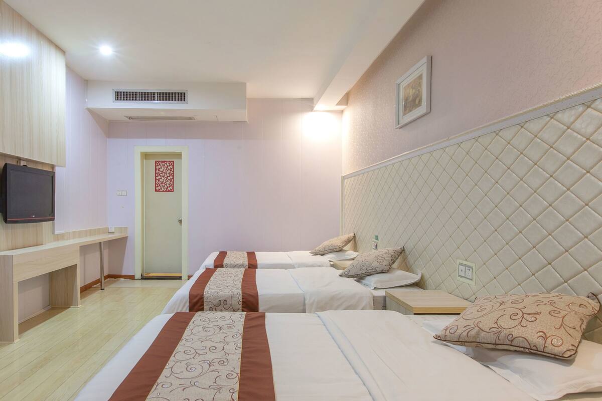 Ein Hotelzimmer mit zwei Betten, einem an der Wand befestigten Fernseher, einer Tür mit dekorativer Verkleidung und einem Deckenlüfter.