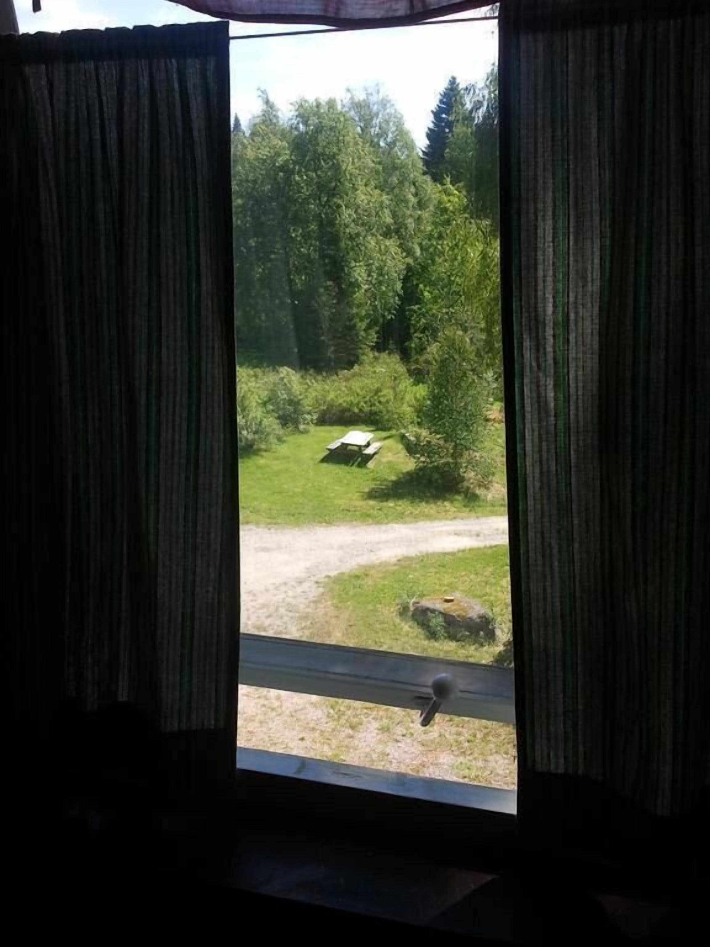 A window with curtains, a view of a grassy area with trees, and a picnic table.