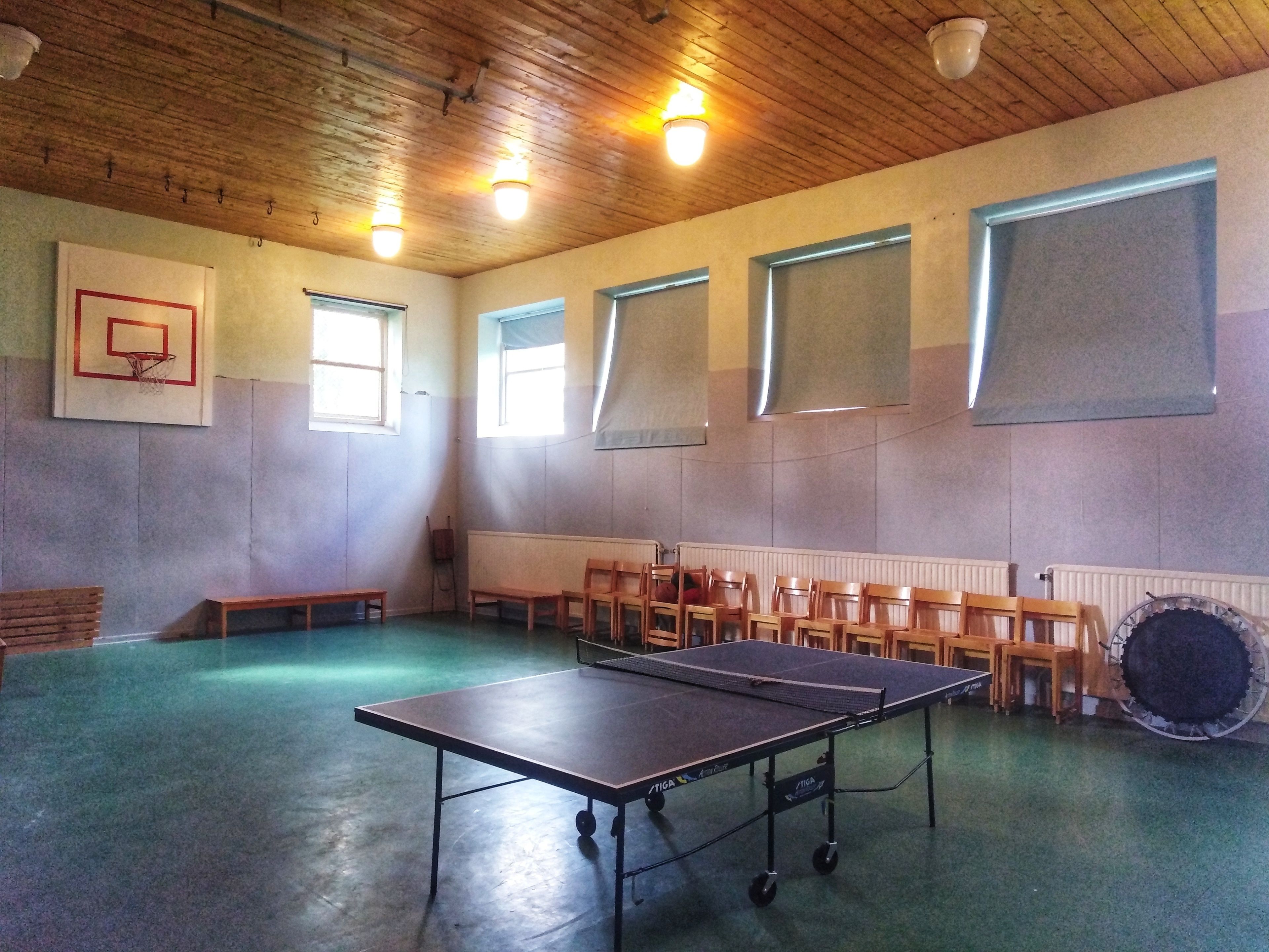 A room with a ping pong table, wooden chairs, a basketball hoop, and windows with blinds.