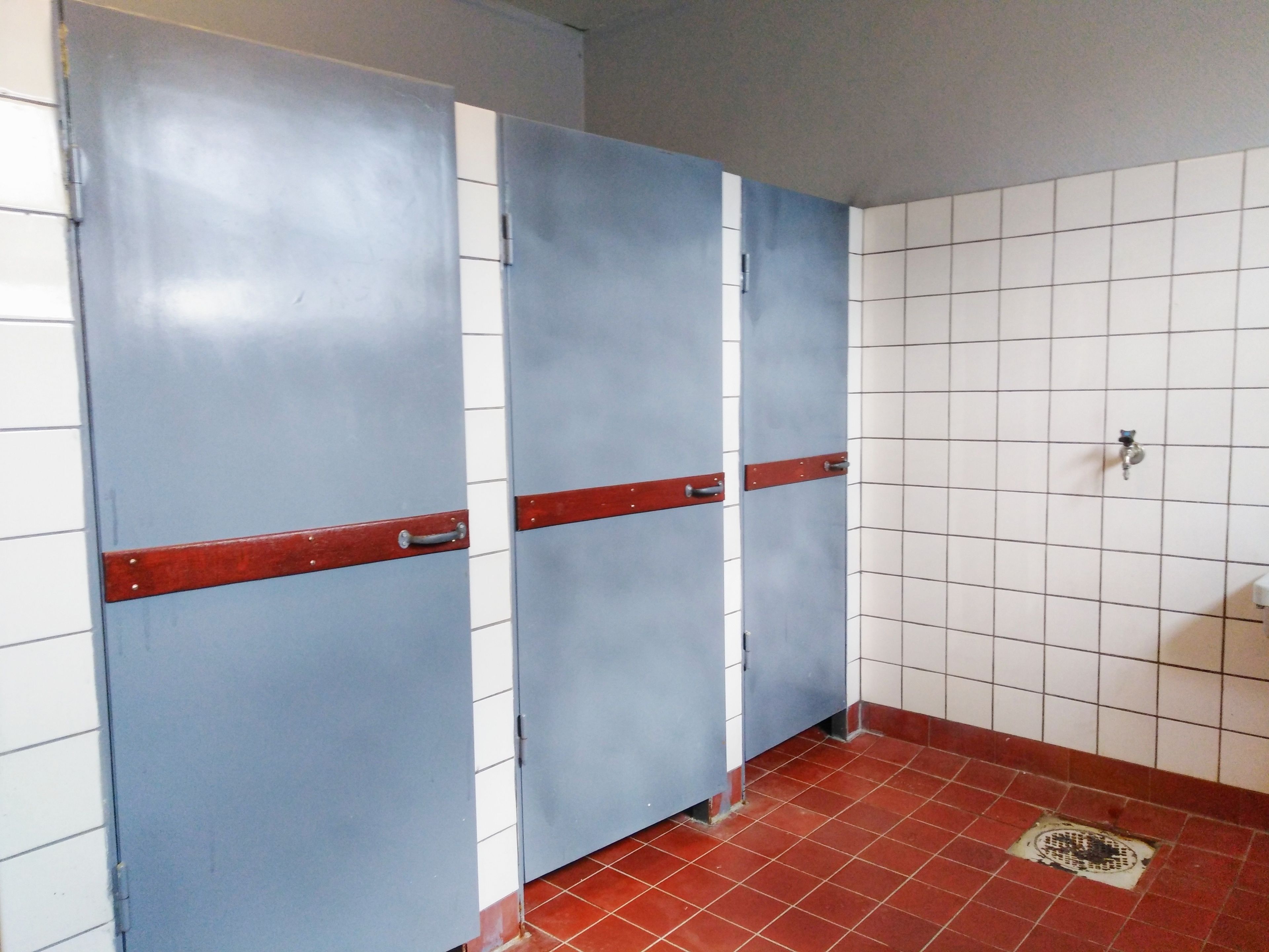 A restroom with three blue stalls, white tiled walls, and a red-tiled floor.