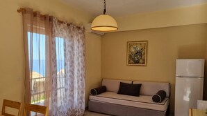 A compact living space with a sofa, a dining table, a refrigerator, and a window with sheer curtains. - Corfu Island Apartment 149 (Corfu)