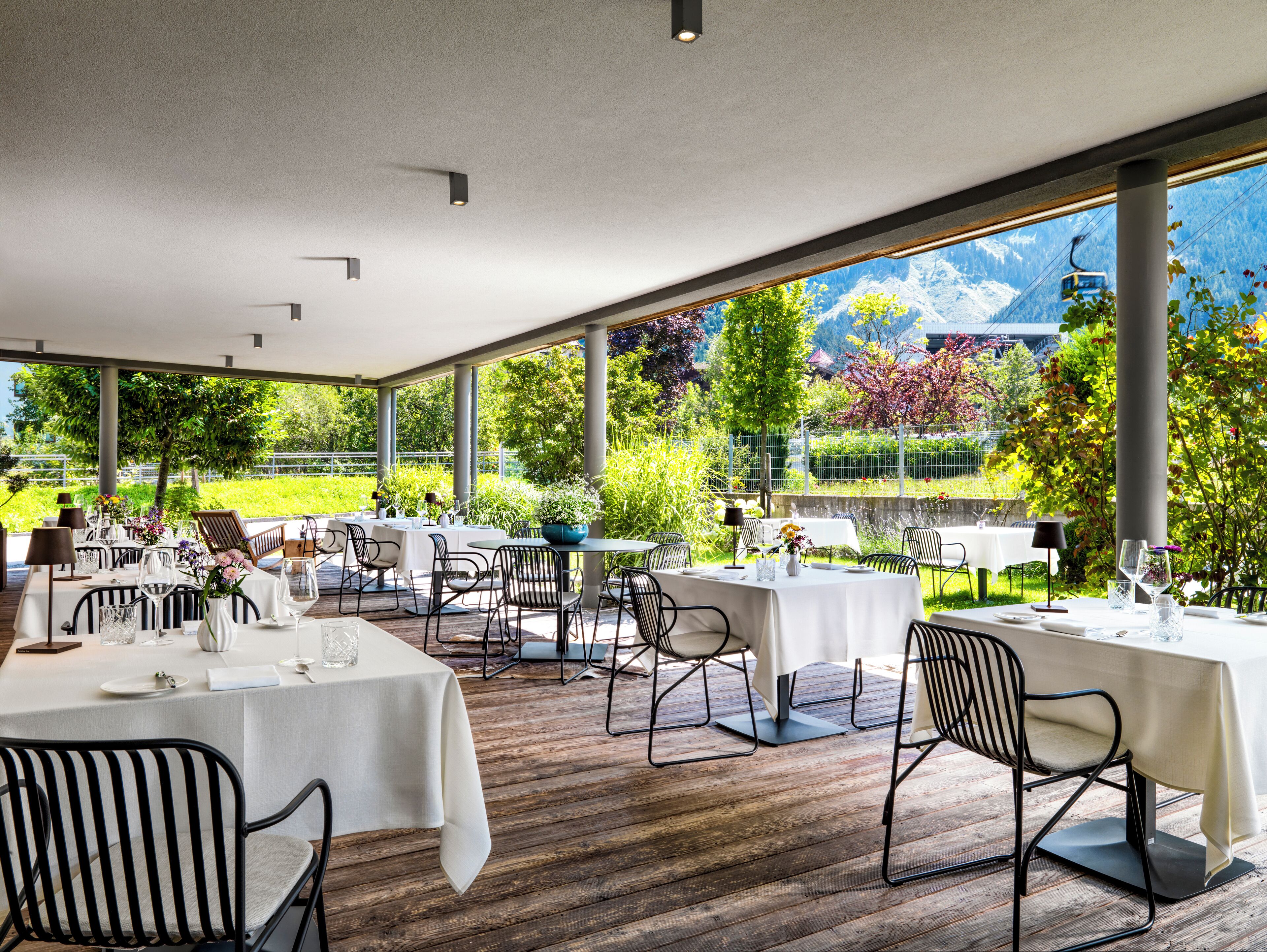 Un área de comedor al aire libre con mesas y sillas, ventanas amplias con vistas a zonas verdes y montañas.