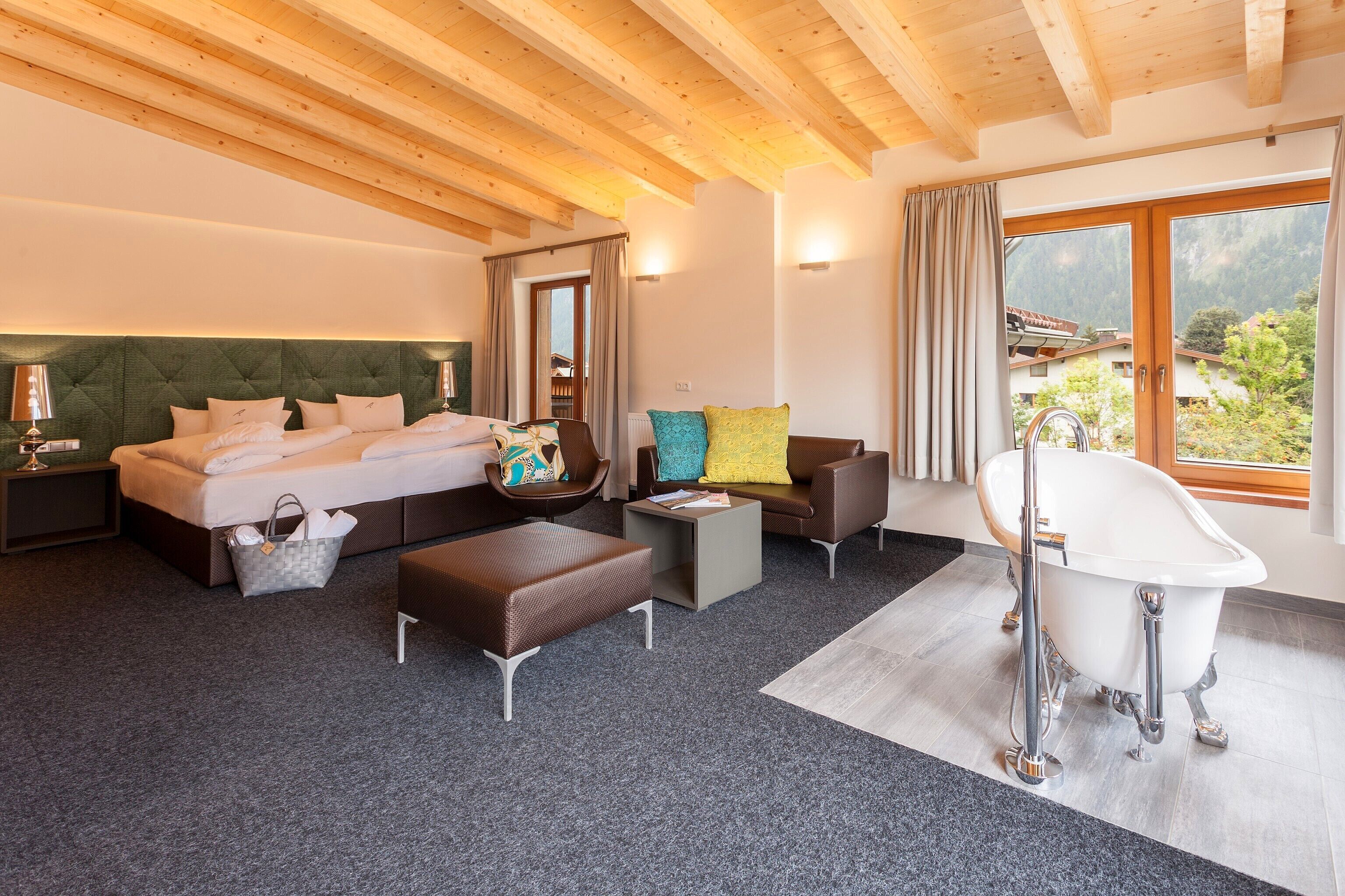A modern hotel room with a large bed, a sitting area with a sofa and armchairs, a small table, a bathtub, and a view of the mountains through the window.