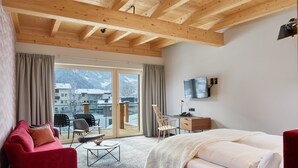 A hotel room with a wooden ceiling, a bed, a desk, a TV, and a view of mountains through the window. - Hubers Boutiquehotel (Mayrhofen)
