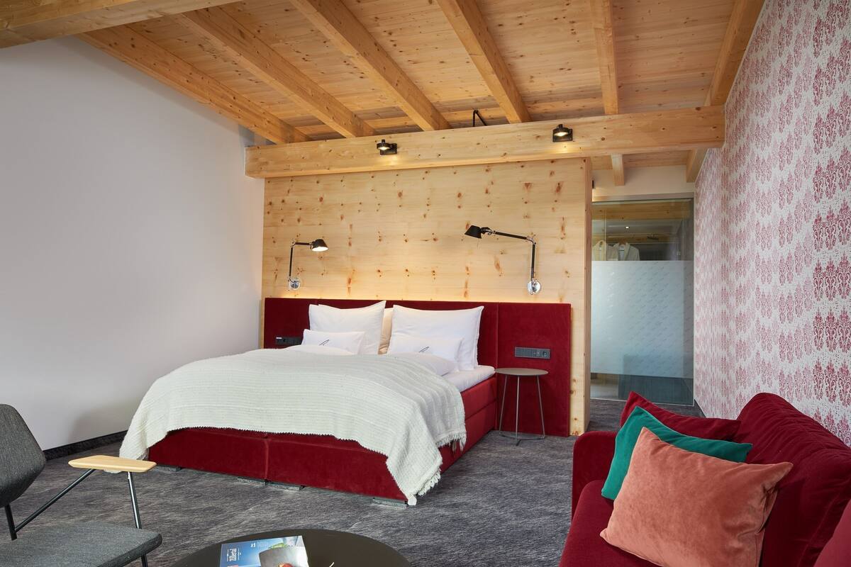 Un dormitorio con techo de madera, una cama con ropa de cama blanca, un cabecero tapizado en rojo, una mesita de noche pequeña, un sillón gris y un sofá rojo con cojines.