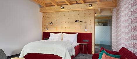 Un dormitorio con techo de madera, una cama con ropa de cama blanca, un cabecero tapizado en rojo, una mesita de noche pequeña, un sillón gris y un sofá rojo con cojines.