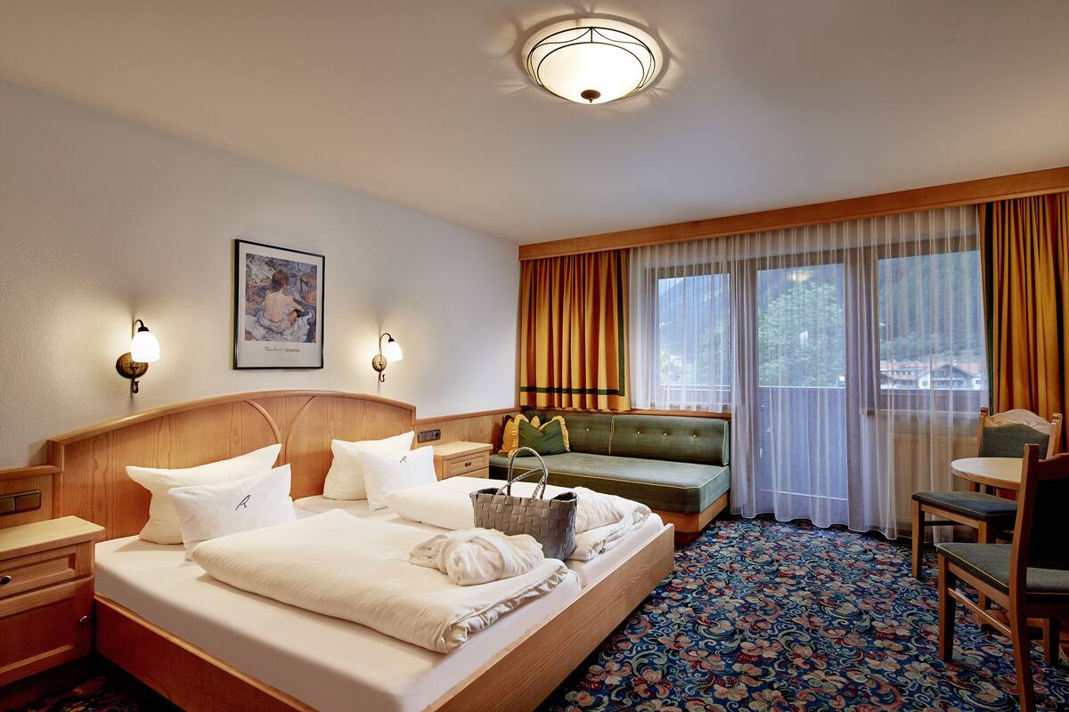 Habitación de hotel con una cama grande, una mesita de noche, una lámpara, un sofá, una silla y un ventanal con cortinas.