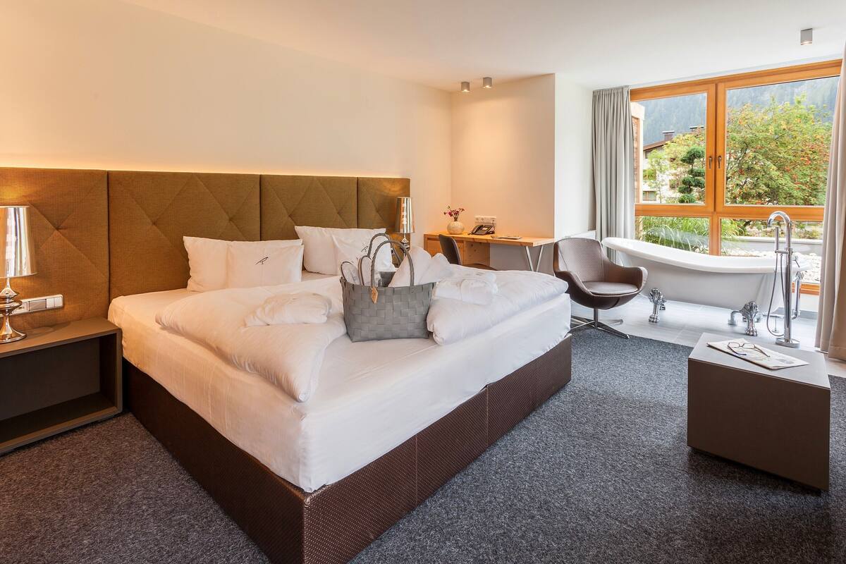 Una habitación de hotel moderna con una cama grande, un escritorio, una silla, una bañera y vistas al exterior.