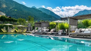 A pool with lounge chairs and umbrellas, surrounded by mountains. - Hubers Boutiquehotel (Mayrhofen)
