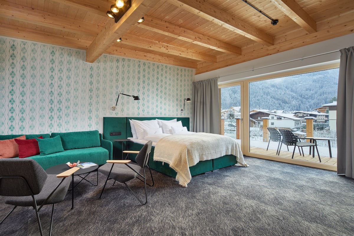 Habitación de hotel con una cama grande, un sofá verde, una mesita y una silla. Tiene un balcón con vistas a las montañas.