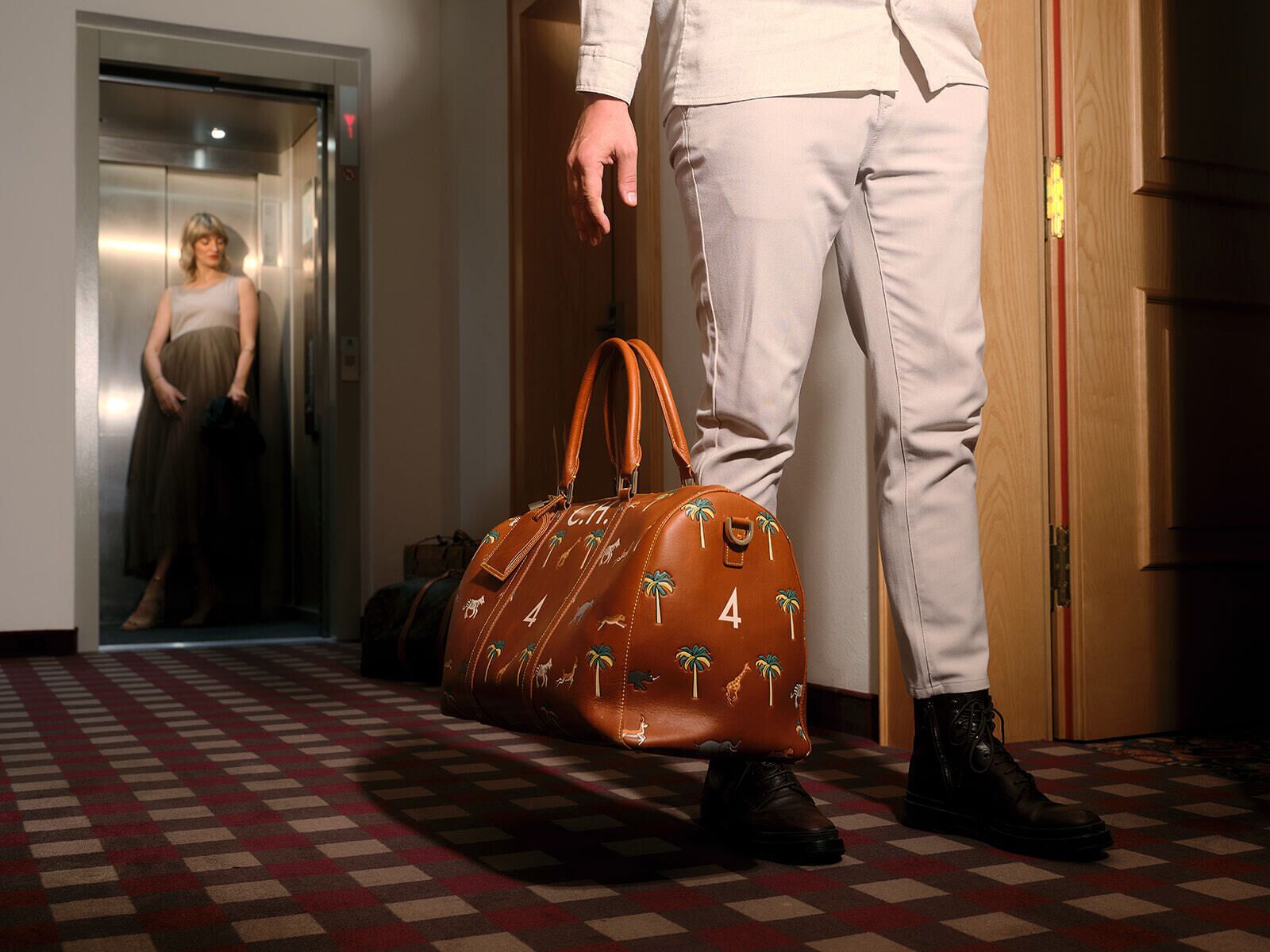 A person standing in a hallway with a checkered floor, wearing white pants and black shoes, holding a brown bag with palm tree designs.