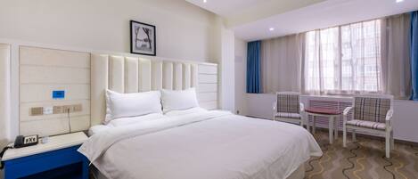A hotel room with a large bed, a bedside table with a phone, a chair, and a window with curtains.