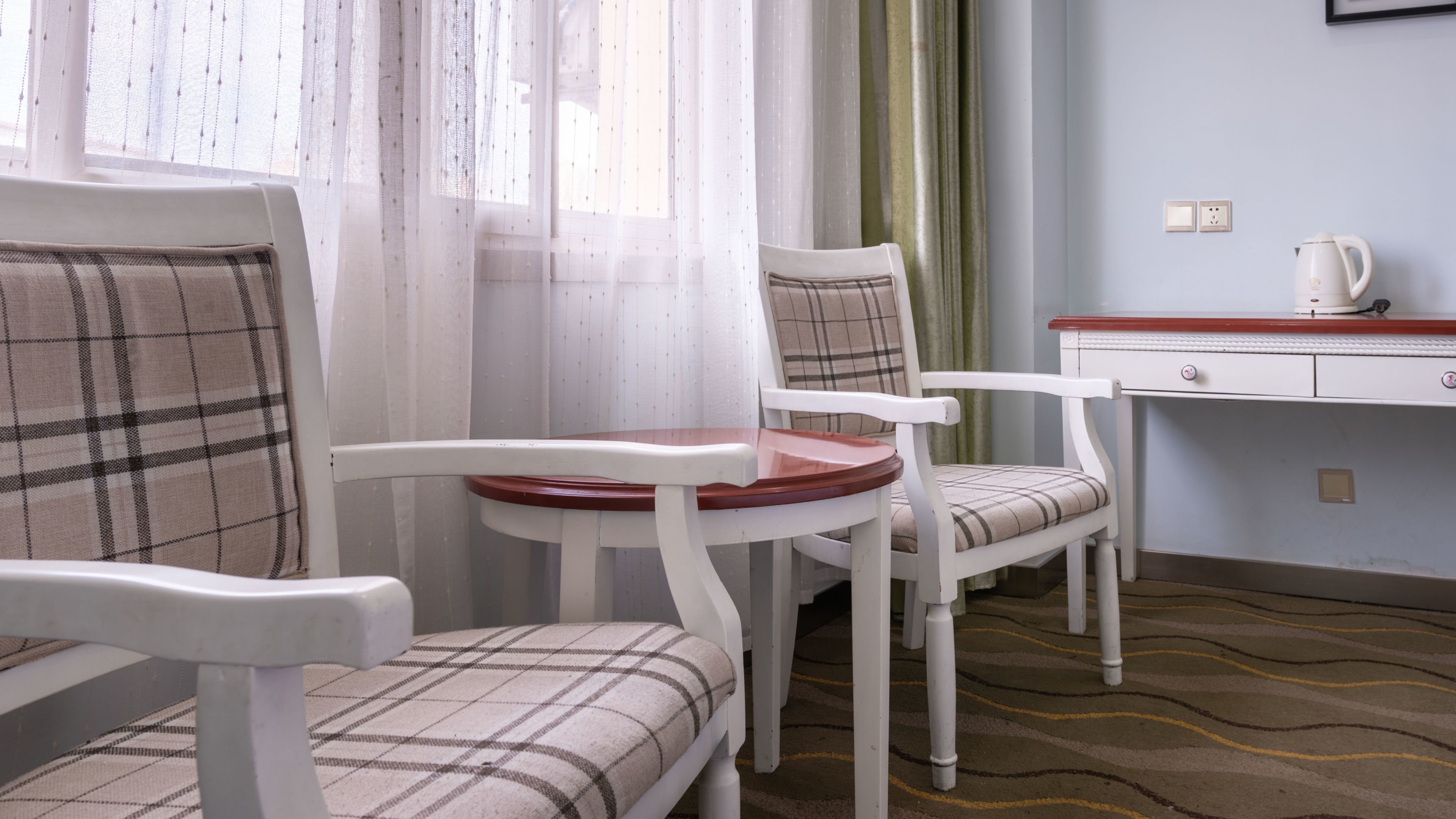 A room with two white chairs with plaid upholstery, a small wooden table, a white dresser with a red top, and a white kettle on the dresser.