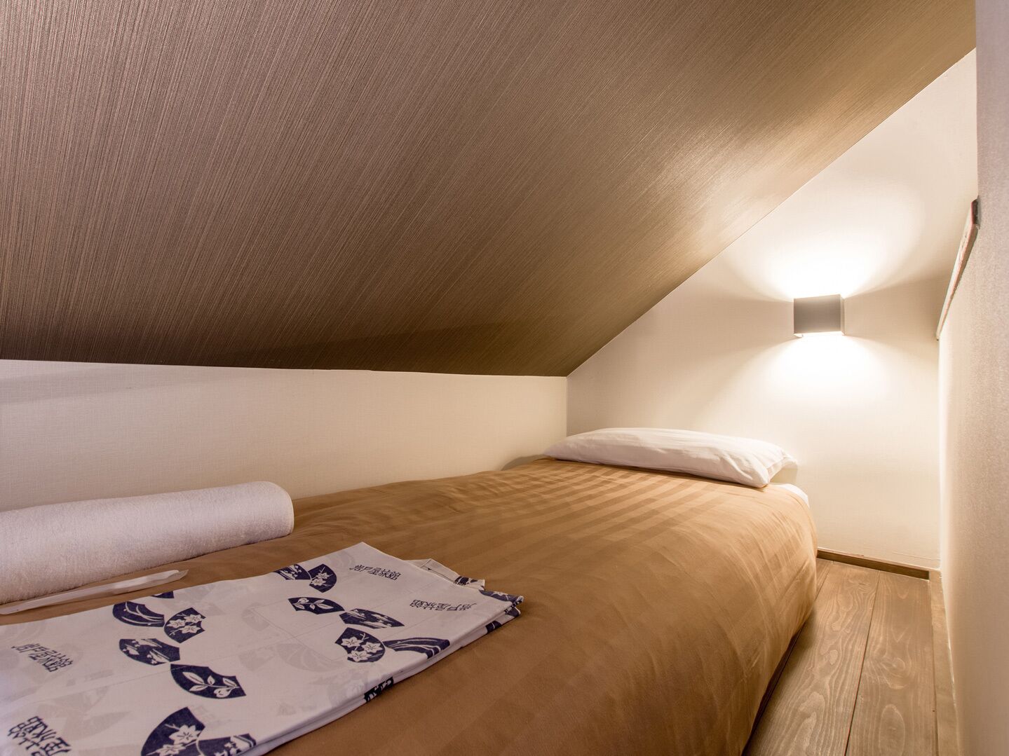 A small, cozy room with a sloped ceiling, a single bed with a white pillow, a neatly folded blanket, and a wall-mounted light fixture.
