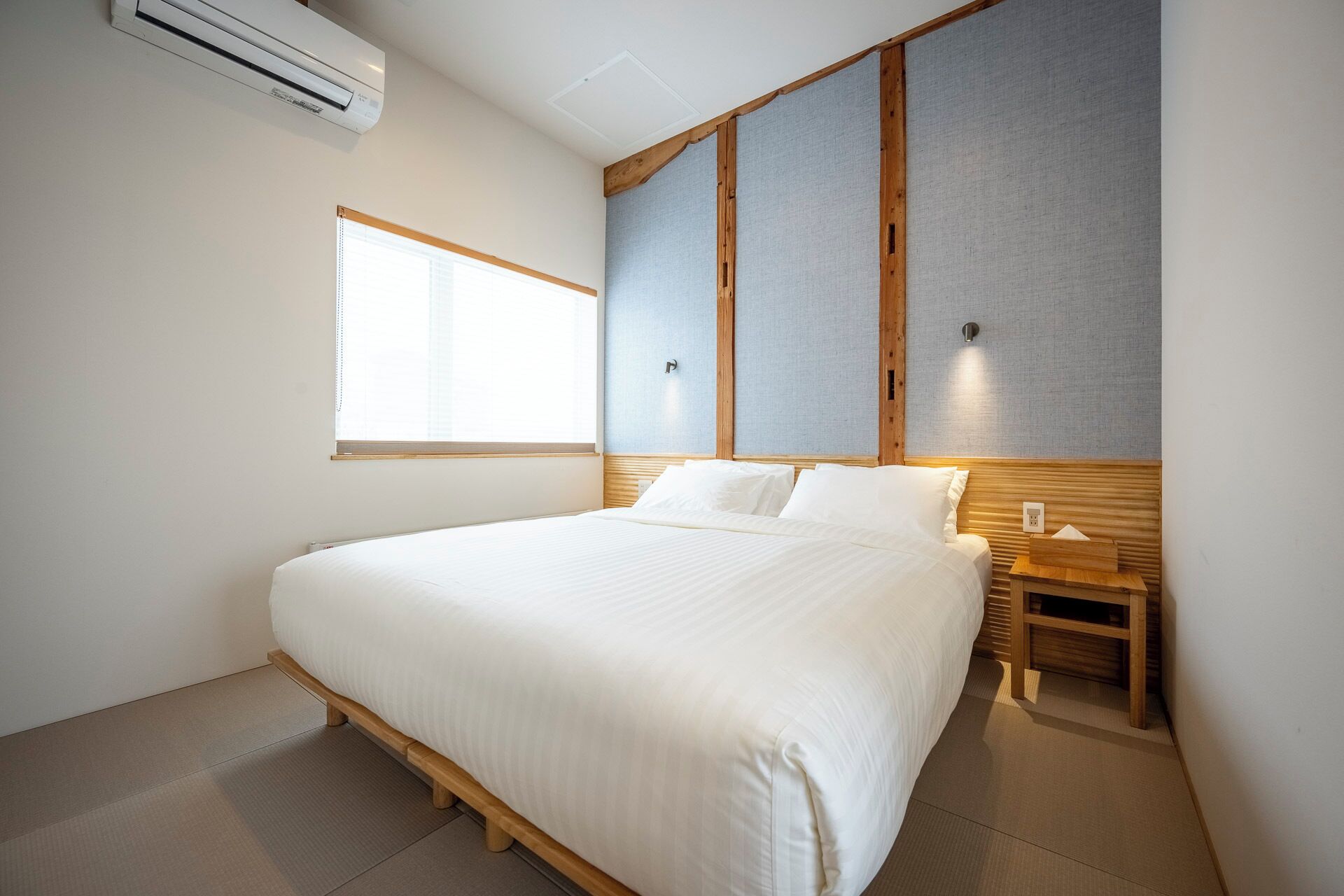 A hotel room with a large bed, a wooden headboard, a bedside table with a lamp, and a window with blinds.