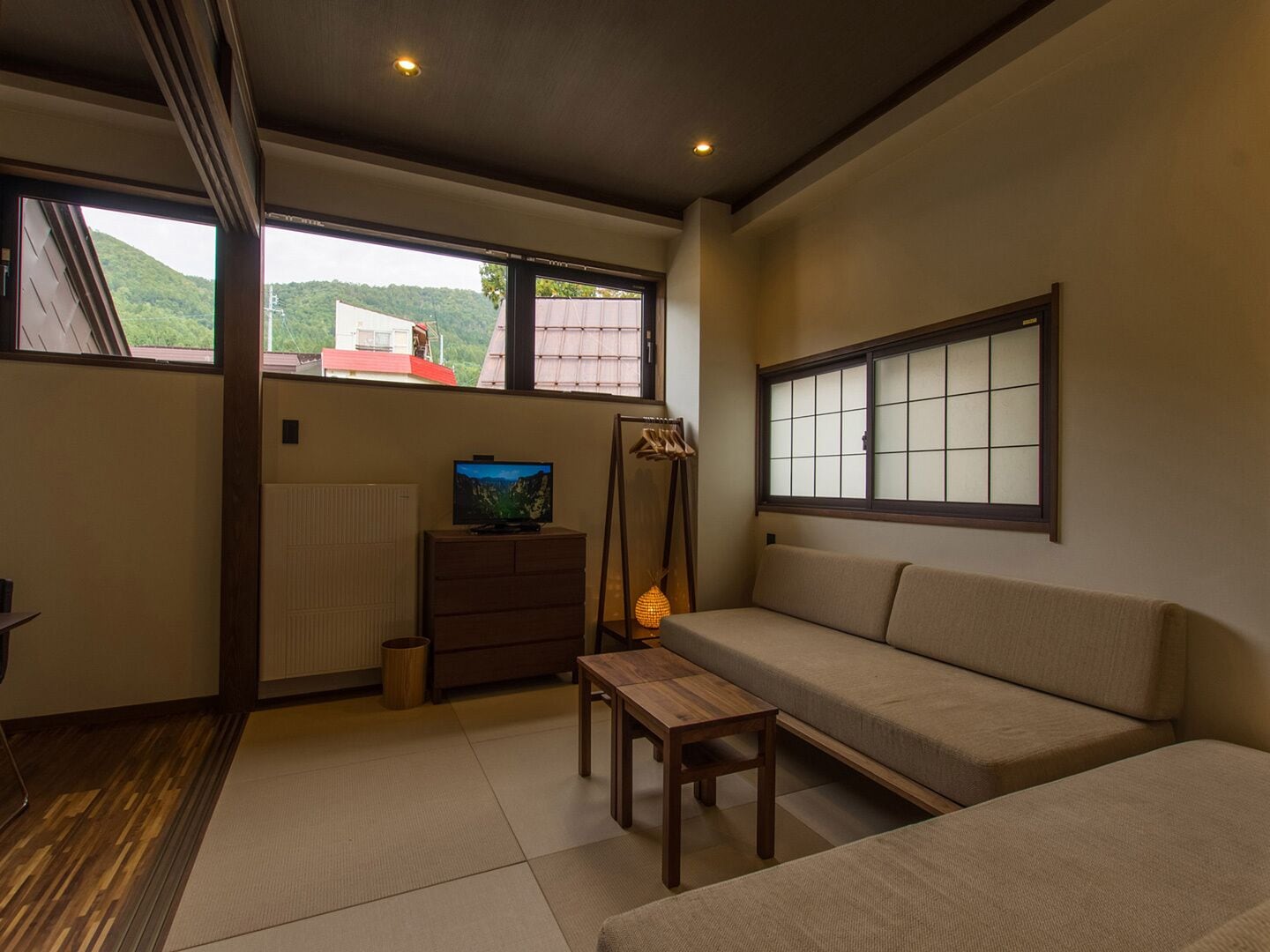 A room with a sofa, a TV on a dresser, a wooden table, and a view of a red-roofed building through large windows.