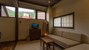 A room with a sofa, a TV on a dresser, a wooden table, and a view of a red-roofed building through large windows.