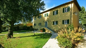 Un edificio giallo a due piani con persiane verdi, un vialetto in pietra e un grande albero sullo sfondo.