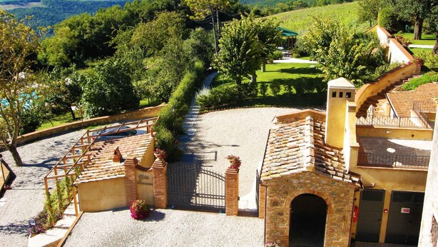Un cortile con un vialetto sterrato, un piccolo edificio con un arco in mattoni e vista su un vigneto e delle colline sullo sfondo.