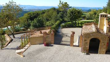 Un patio con un sendero de grava que conduce a una entrada con puerta, rodeado de abundante vegetación y vistas a colinas distantes.