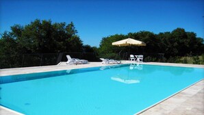 Una piscina rettangolare con acqua cristallina, circondata da un solarium piastrellato. Si vedono lettini da spiaggia e un ombrellone che offre ombra.