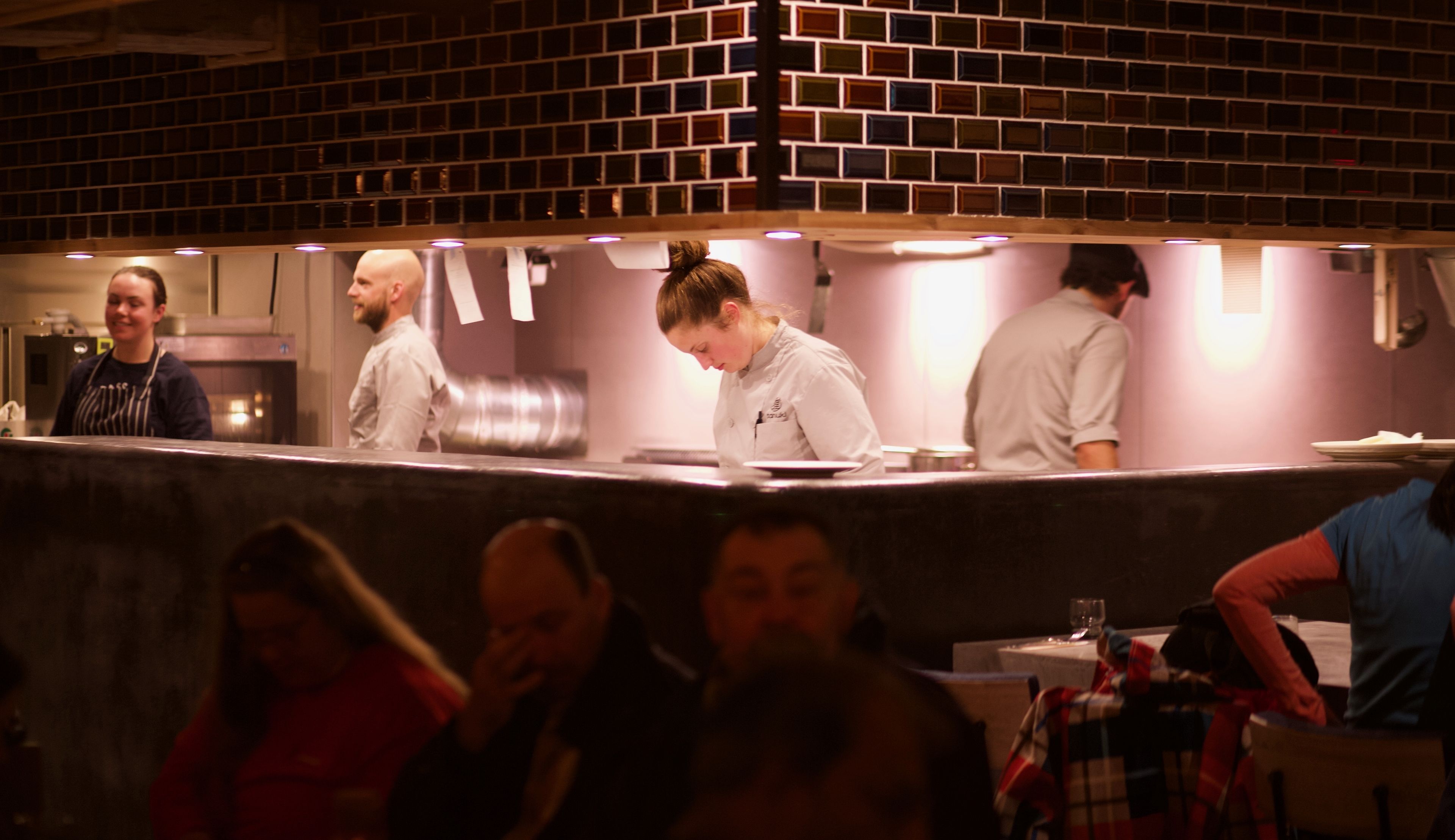 A restaurant interior with a brick wall, a long counter, and a group of people seated at tables.