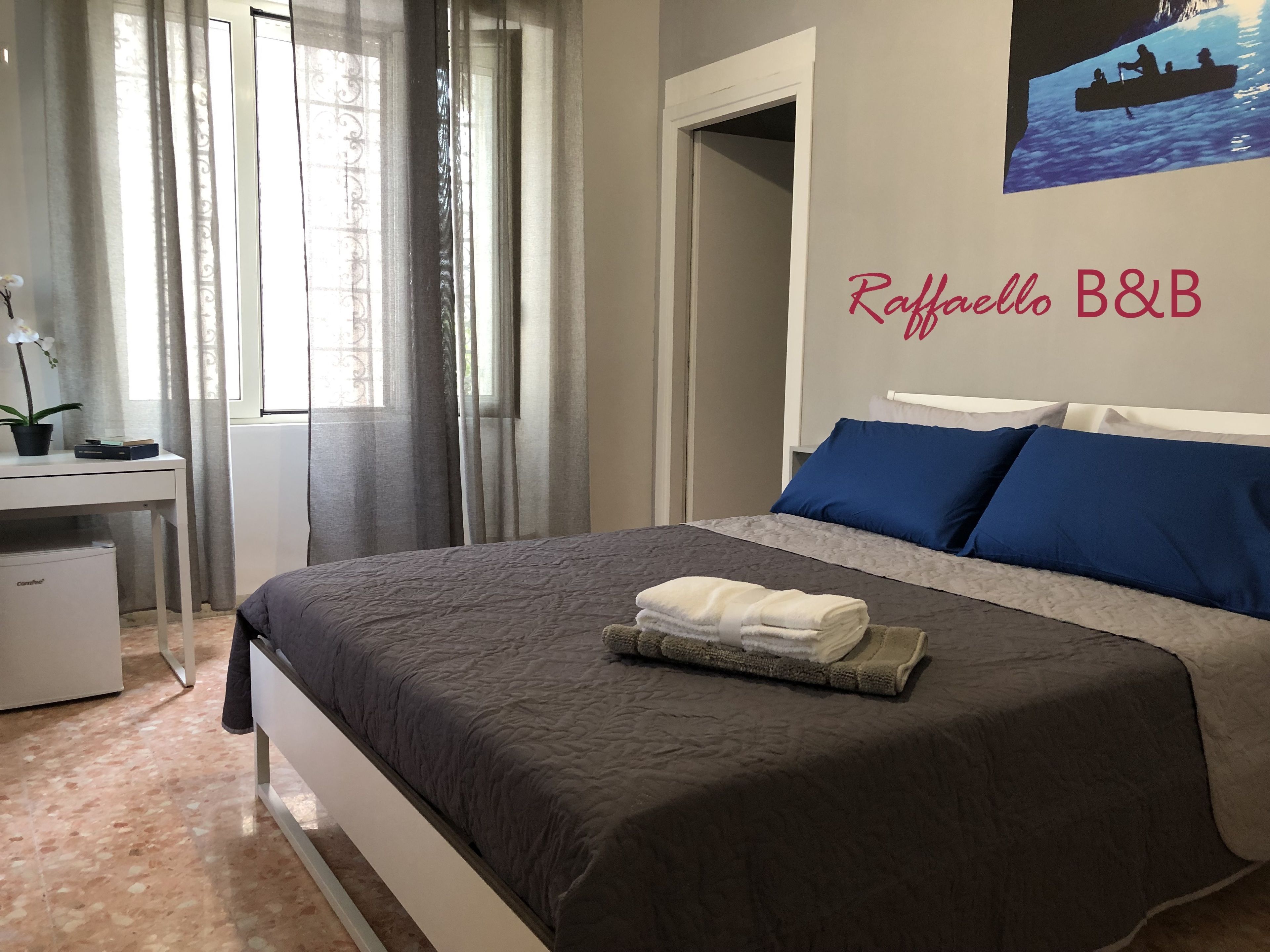 Una cama bien hecha con una toalla doblada, un escritorio con una planta en maceta, y una pared con un cuadro y texto.