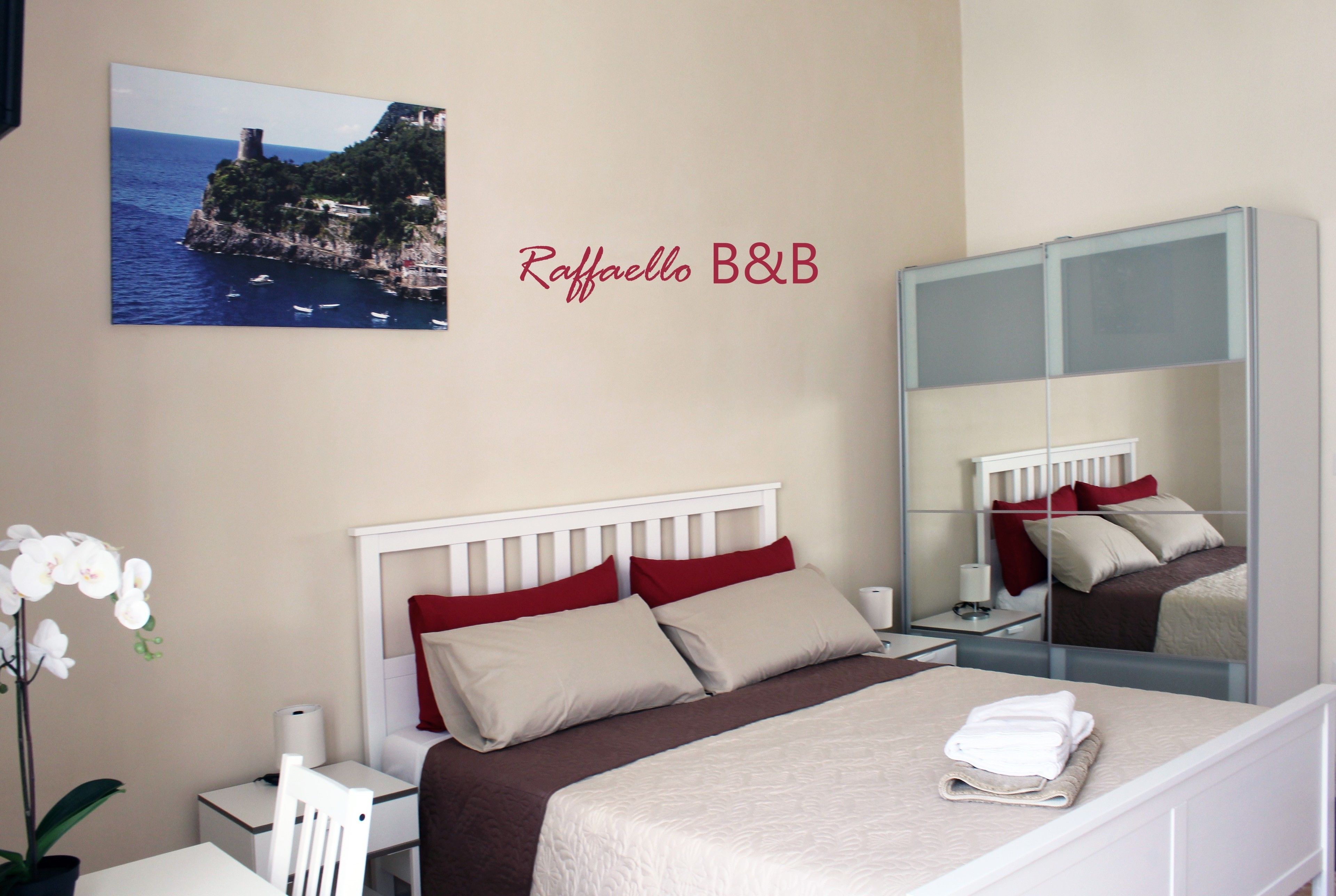 Et hotelværelse med en seng, et natbord, et spejl, et billede af et kystlandskab og et skilt med teksten "Raffaello B&B".
