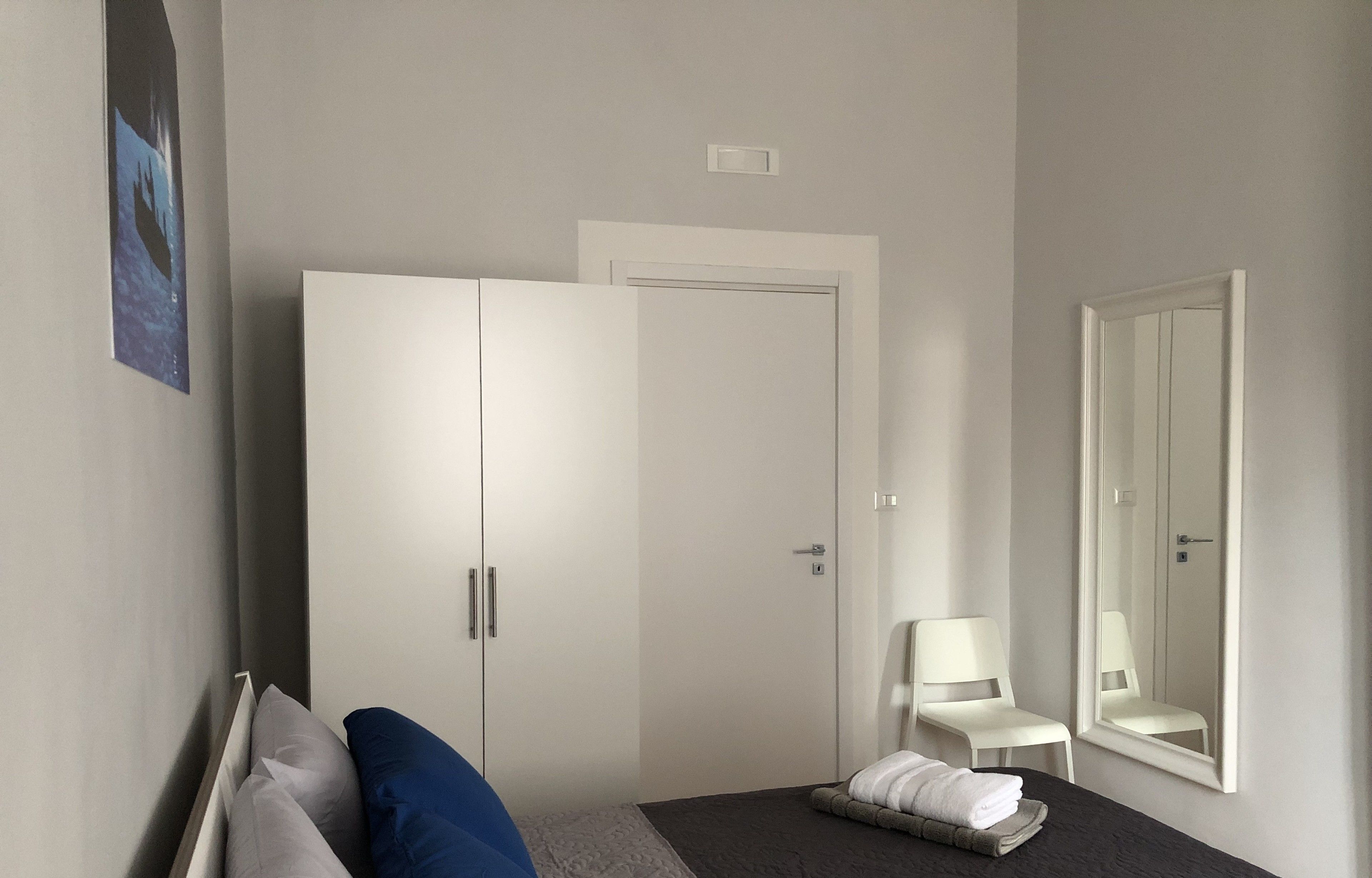 A hotel room with a bed, a wardrobe, a chair, a mirror, and a small table with a towel.