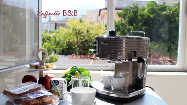 A breakfast setup with a coffee maker, cups, and sandwiches on a table by a window. - B & B Raffaello (Terzigno)