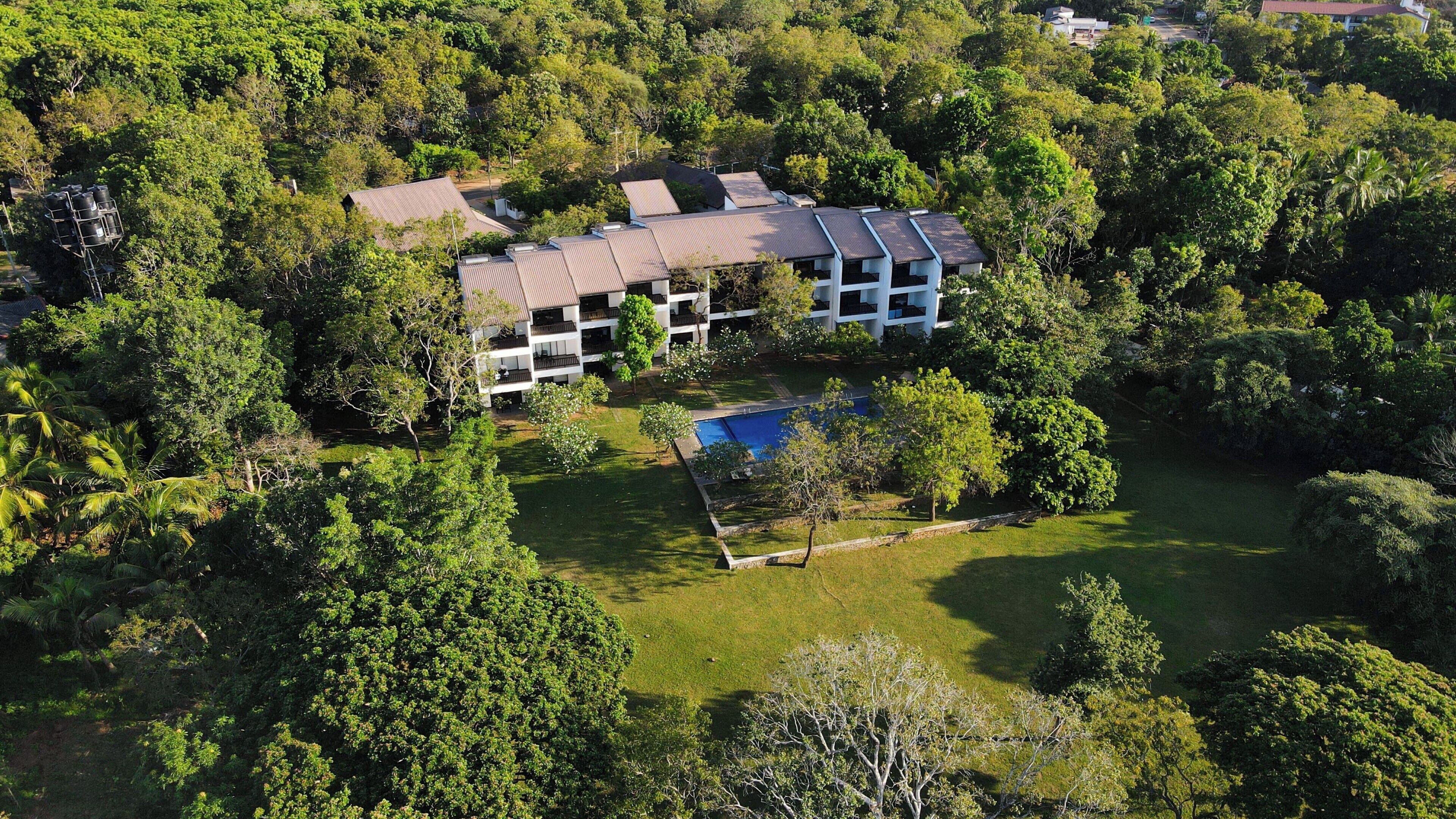 A multi-story building surrounded by lush greenery, with a swimming pool and a well-maintained lawn.