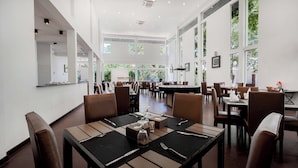 Un restaurant spacieux, doté de hauts plafonds et de grandes fenêtres, qui offre une vue sur la verdure environnante. Les tables sont dressées avec une vaisselle simple.