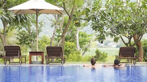Zona de piscina con una persona relajándose en el agua, rodeada de árboles y mobiliario exterior.