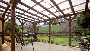 A covered patio area with a grill, chairs, and a view of a garden and houses in the background.