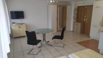A compact hotel room with a bed, a small round table with two chairs, a television mounted on the wall, and a kitchenette area with cabinets and a refrigerator.